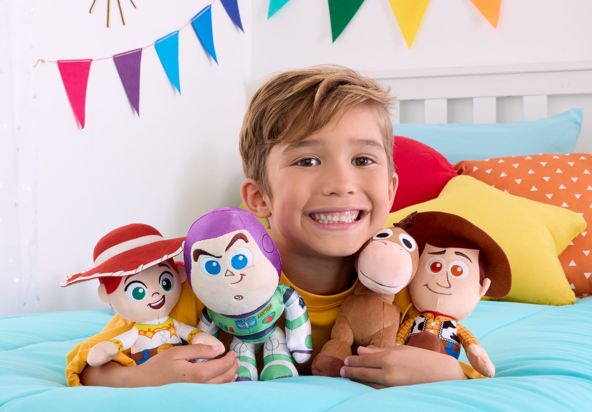 Child holding Buzz Lightyear, Woody, and other plush toys on a bed with colorful pillows and flags.